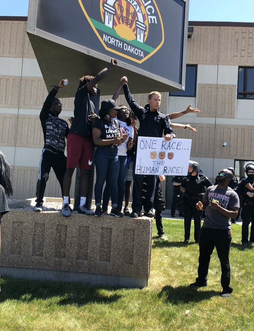 Officer holding hands with protest organizers in North Dakota & a sign ‘We are one race... The HUMAN race.